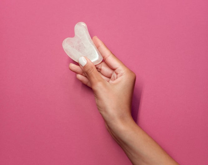 una mano sosteniendo una herramienta de gua sha con un fondo rosa pastel