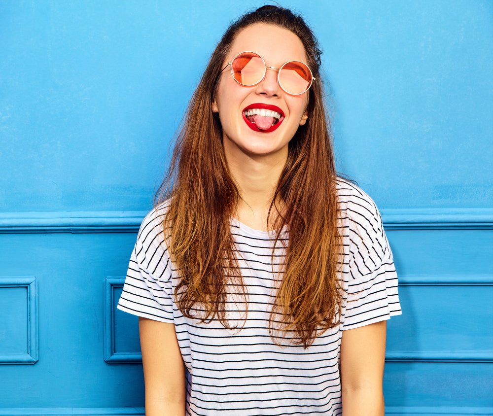 modelo con los labios pintados de rojo sonriendo y sacando la lengua con lentes naranjas en un fondo azul